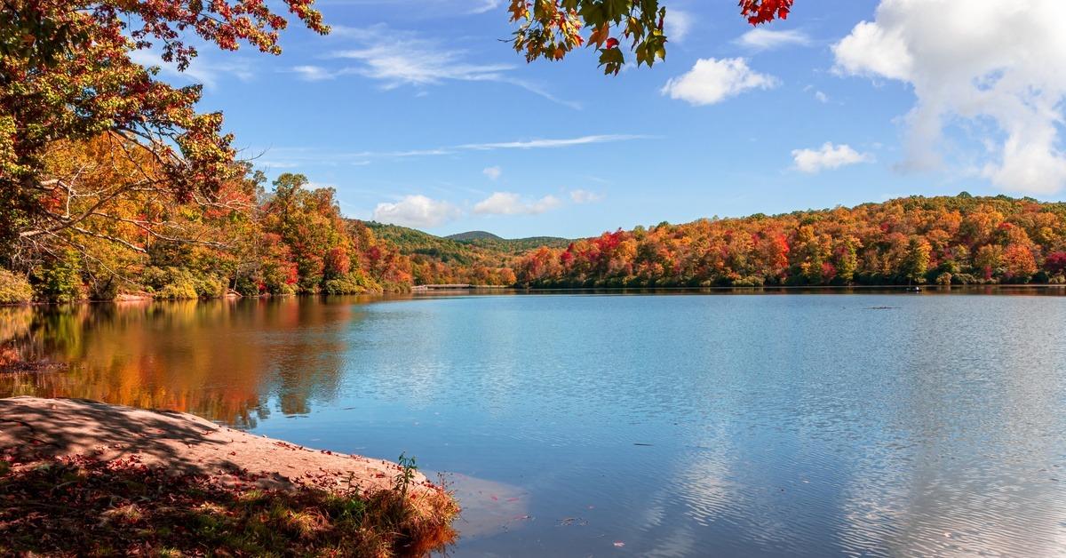 Trees with red, orange, and yellow leaves line a large lake. The blue sky contains white clouds.