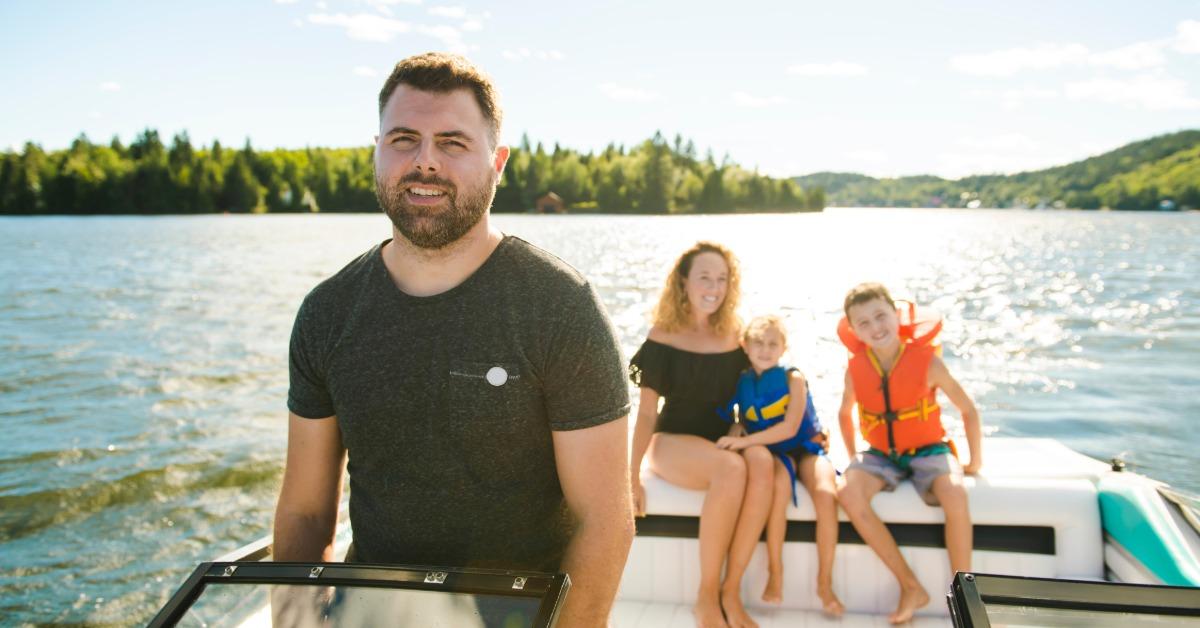 A man is standing up, driving a boat while a mom and two young boys sit on the back. The boys are wearing life vests.