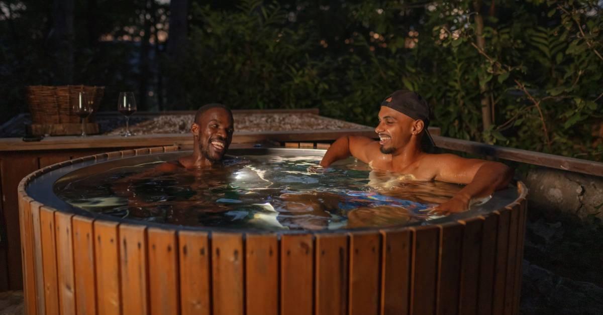 Two men sitting in a hot tub together as the sun sets behind the trees. The hot tub is surrounded by wood panels.