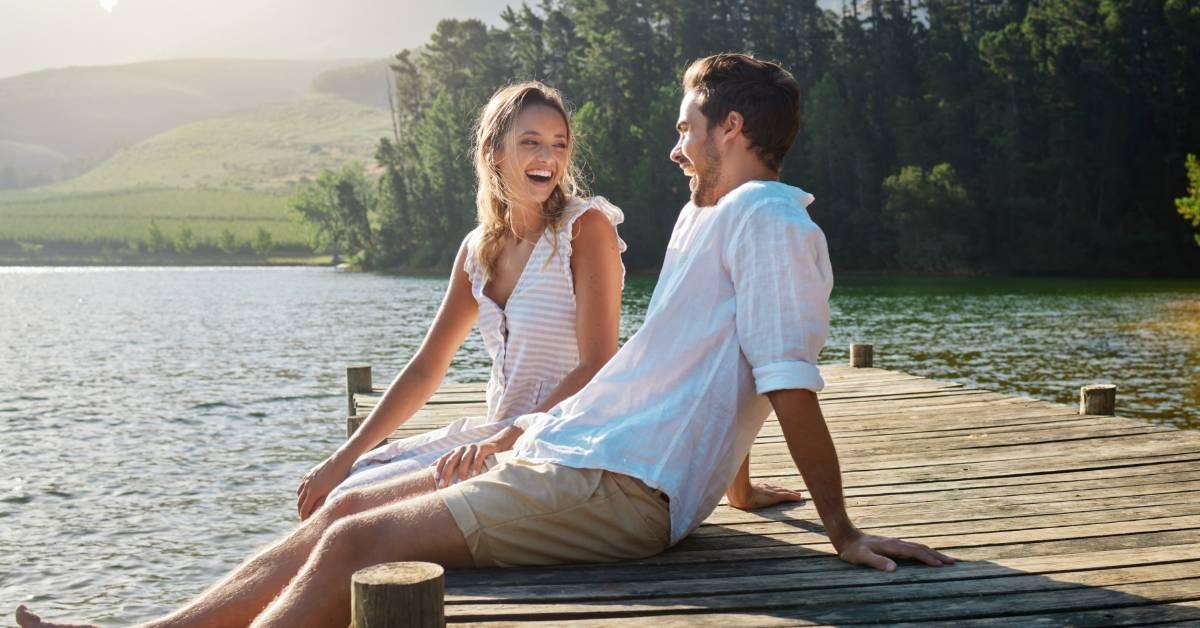 A man and a woman sitting on a deck that stretches out into a lake. They are both looking at each other and laughing.
