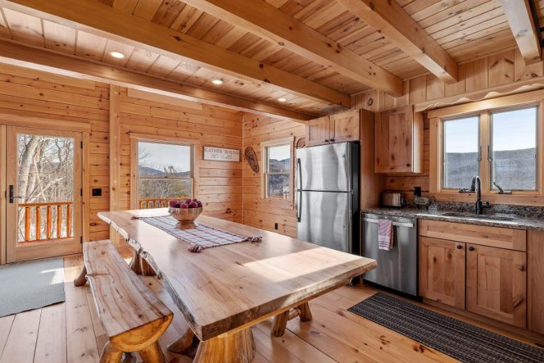 A log cabin interior with a kitchen and dining table. It features wood walls, multiple windows, and mountain views.