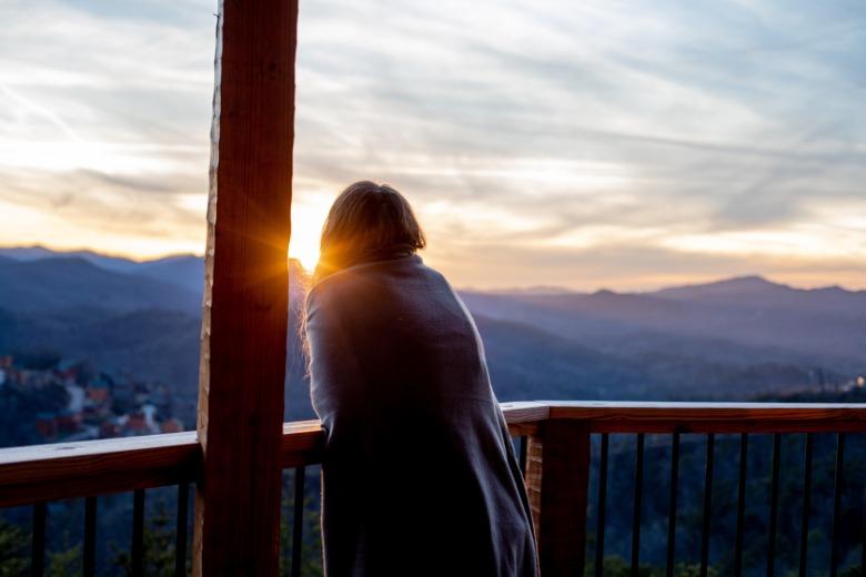 A young woman stands on a cabin deck looking out toward a mountain landscape with trees and distant peaks.
