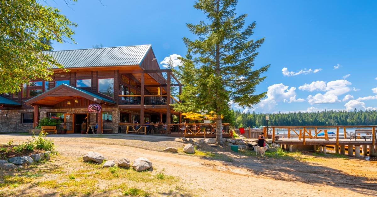 A lakeside lodge sitting beside a sandy beach and wooden dock, with calm water and tall trees under a blue sky.