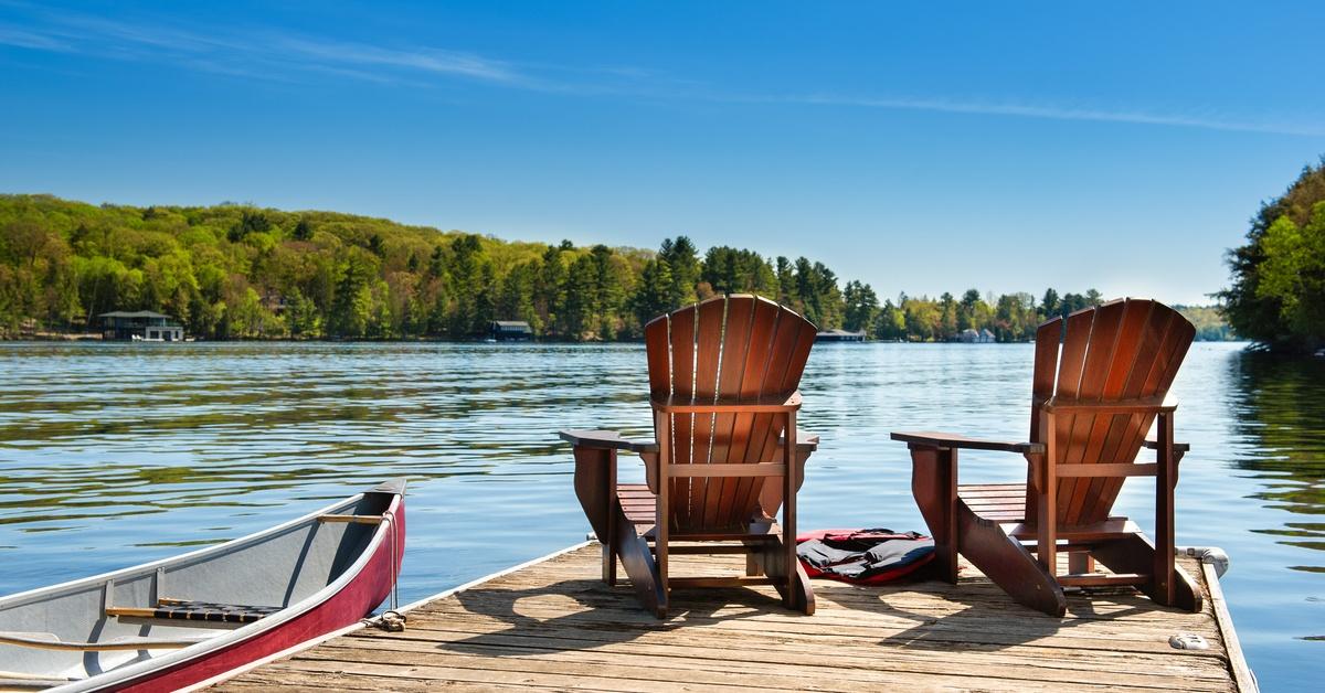 Two wooden chairs sitting on a dock beside a red canoe, facing a calm lake with a tree-covered shoreline.