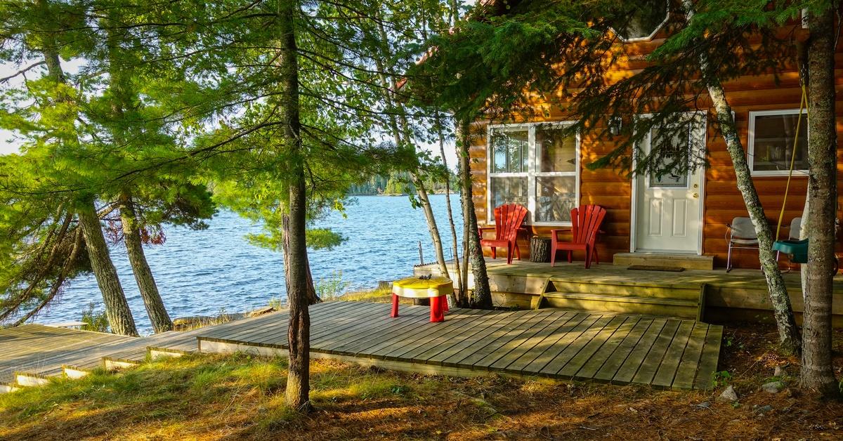 A log cabin sitting beside a lake, with red chairs on a wooden deck and tall trees surrounding the porch.